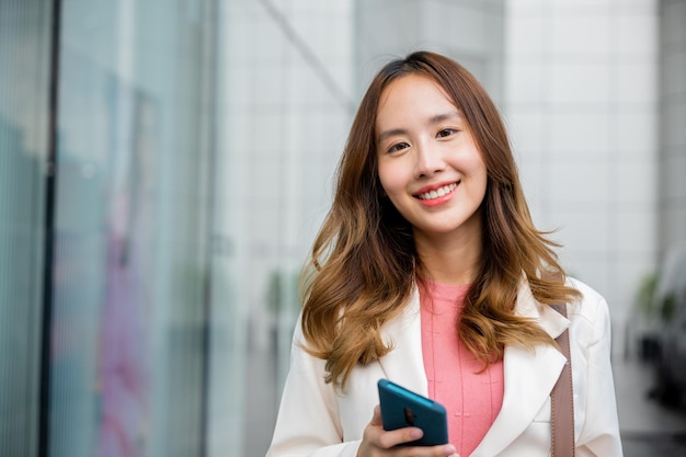 Empresária asiática, enviando mensagens de texto para smartphone, viajando para o trabalho, ela andando perto de seu prédio de escritórios, Mulher de negócios feliz sorrindo usando telefone celular caminhando ao ar livre na rua da cidade urbana olhando para a câmera