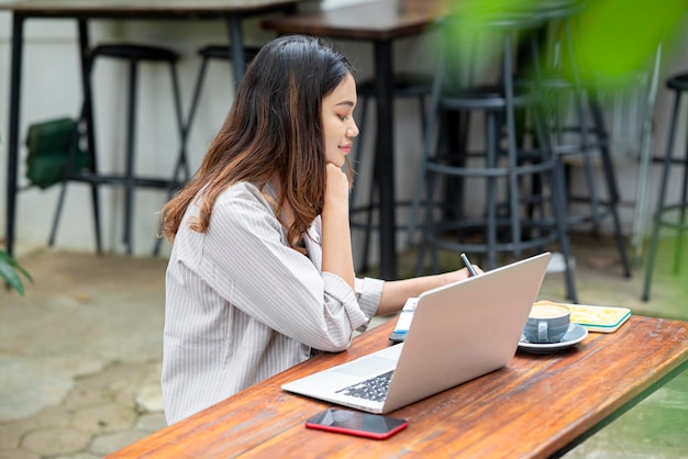 Empresária asiática escreve em um caderno com um laptop de celular e uma xícara de café na mesa