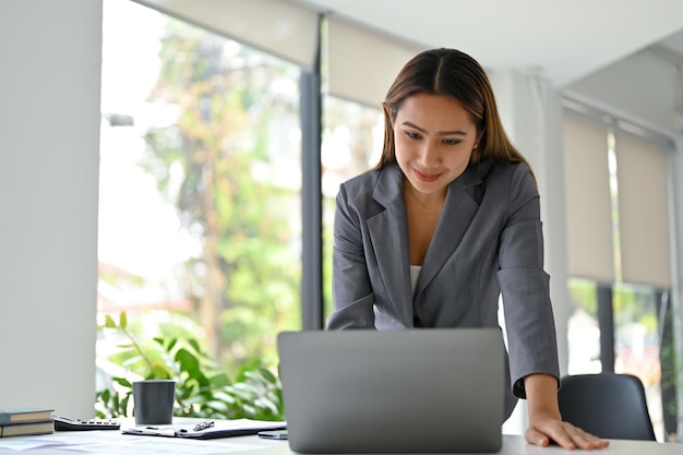 Empresária asiática inteligente, apoiando-se na mesa e trabalhando em seu projeto de negócios no laptop