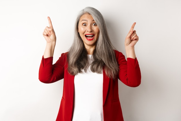 Empresaria asiática madura con cabello gris, vistiendo chaqueta roja y apuntando con el dedo hacia arriba, sonriendo sorprendido, mostrando oferta promocional, fondo blanco.