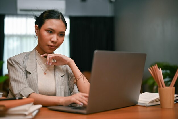 Empresaria asiática milenaria profesional y segura que trabaja en sus tareas en una computadora portátil