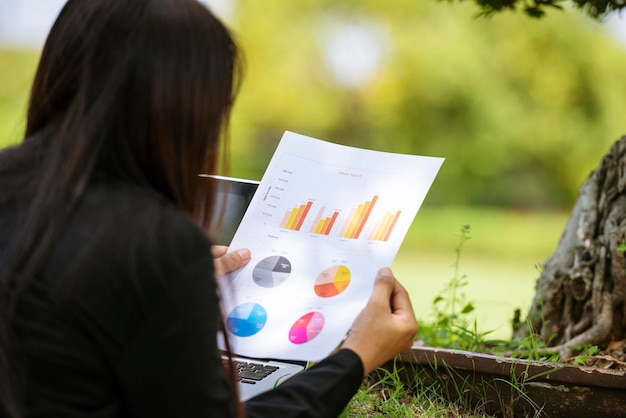 Empresaria asiática mujer de negocios leyendo gráfico de negocios analizar gráfico usando una computadora portátil en un jardín verde en casa Trabajo independiente en la oficina en casa distancia social nuevo estilo de vida normal en un café abierto parque verde