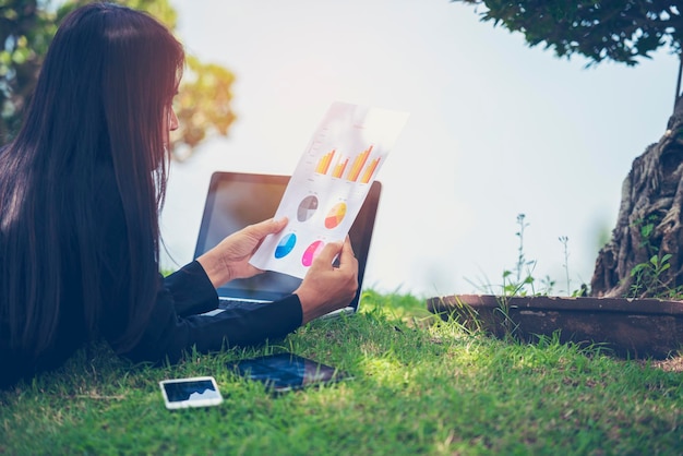 Empresaria asiática mujer de negocios leyendo gráfico de negocios analizar gráfico usando una computadora portátil en un jardín verde en casa Trabajo independiente en la oficina en casa distancia social nuevo estilo de vida normal en un café abierto parque verde