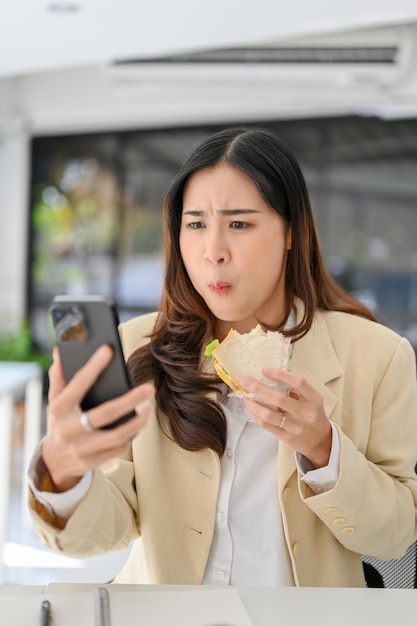 Empresária asiática ocupada, com fome e sobrecarregada, comendo um sanduíche enquanto usa o telefone