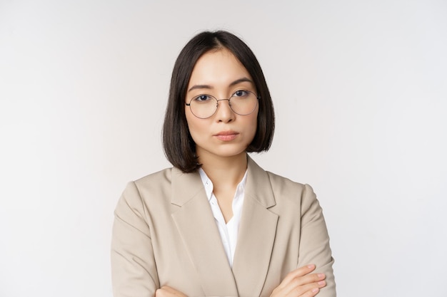Empresária asiática profissional em óculos parecendo confiante na câmera em pé em pose de poder contra fundo branco