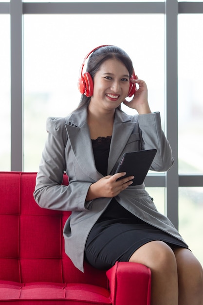 Empresaria asiática soñadora en auriculares sentado en el sofá en el pasillo del centro de negocios y disfrutando de canciones con pose fácil durante el descanso en el trabajo