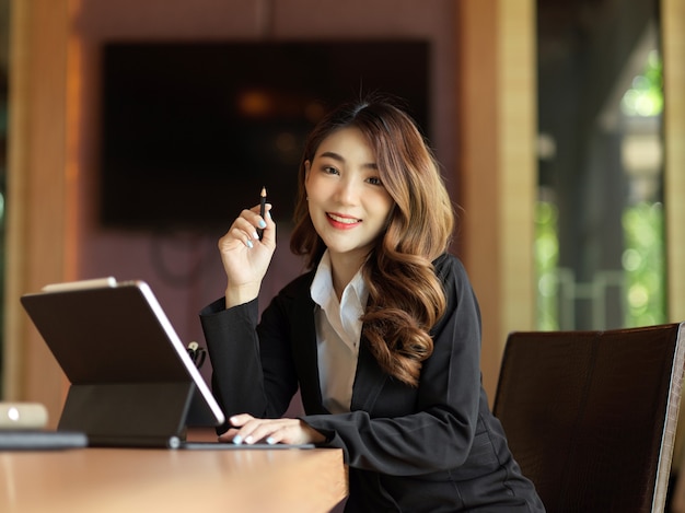 Empresaria asiática sonriendo con confianza trabajando con su tableta inteligente sentada en su escritorio de oficina