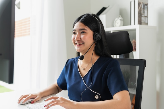 Empresaria asiática sonriente que trabaja en el escritorio de oficina con las auriculares y la computadora.