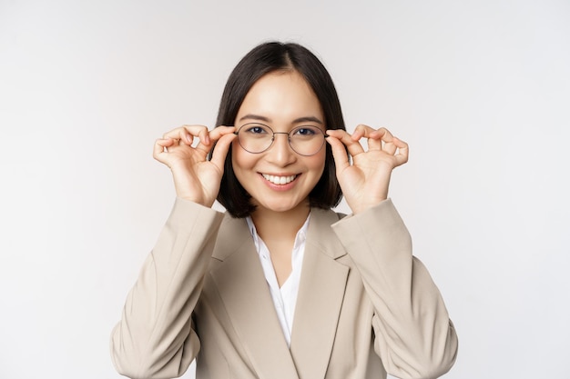 Empresária asiática sorridente tentando novos óculos usando óculos em pé de terno sobre fundo branco