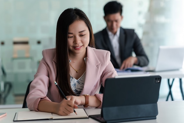 Empresaria asiática sosteniendo un lápiz sonriendo feliz en el trabajo usando una tableta en la oficina la parte posterior está borrosa.