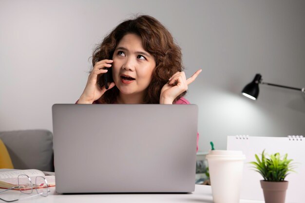 Empresária asiática trabalha em casa com laptop e falando no smartphone e sorrindo. Mulher trabalhando no computador e usando o celular enquanto está sentado em casa