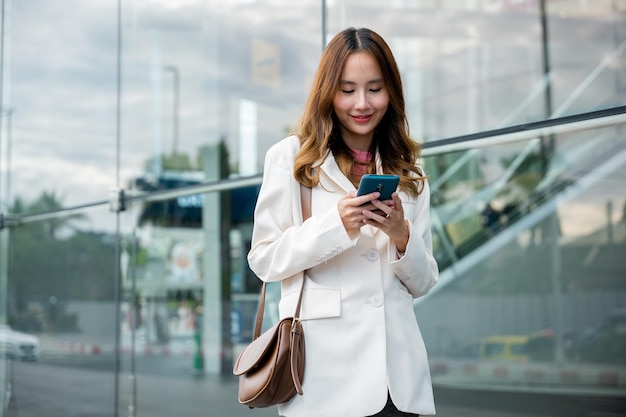 Empresária asiática trabalhando com telefone celular em frente ao prédio da rua perto do escritório, Retrato de uma mulher de negócios bem-sucedida sorrindo segurando o bate-papo do aplicativo de uso do smartphone on-line pela manhã