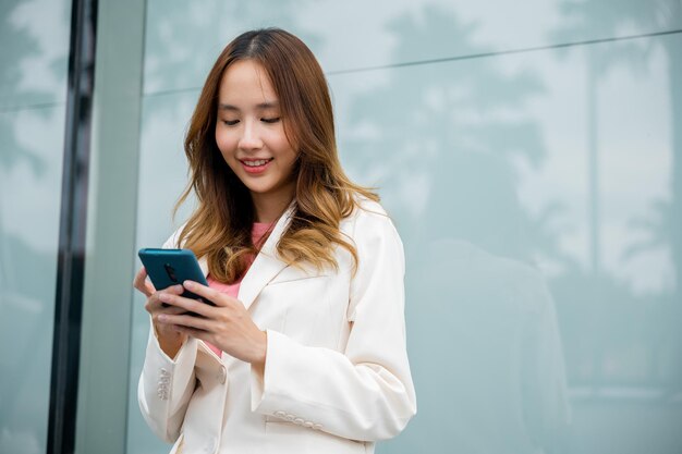 Empresária asiática trabalhando com telefone celular em frente ao prédio da rua perto do escritório, Retrato de uma mulher de negócios bem-sucedida sorrindo segurando o bate-papo do aplicativo de uso do smartphone on-line pela manhã