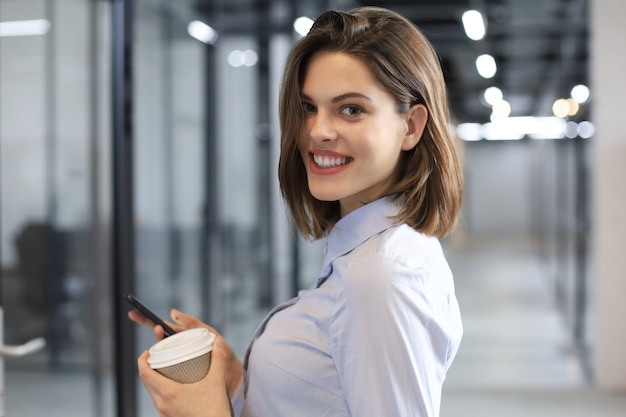 Empresaria caminando por el pasillo de la oficina con vaso de papel y smartphone.