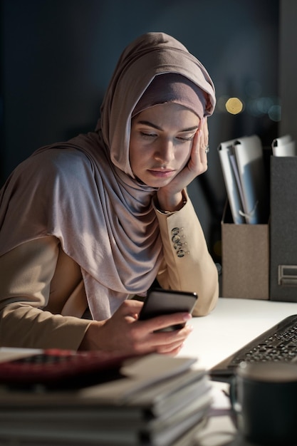 Empresaria cansada en hijab mirando a través de mensajes en el teléfono inteligente