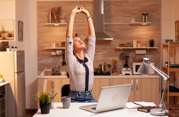 Empresária cansada se espreguiçando enquanto trabalhava horas extras na cozinha de casa. Funcionário que usa tecnologia moderna à meia-noite fazendo horas extras para trabalho, negócios, ocupado, carreira, rede, estilo de vida, sem fio.