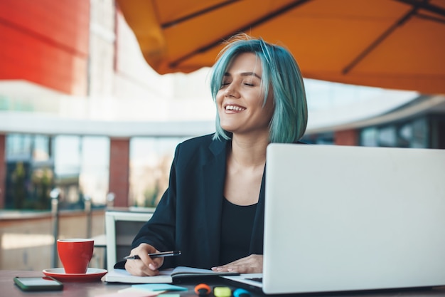 Empresaria caucásica de pelo azul está sonriendo en una cafetería bebiendo un té y trabajando en la computadora portátil