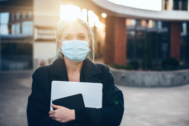 Empresaria caucásica de pelo azul con una máscara protectora mientras sostiene una computadora portátil y posando afuera