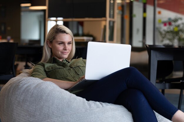 Empresaria caucásica usando una computadora portátil sentada en un sillón en una oficina creativa. tecnología y distanciamiento social en el lugar de trabajo de oficinas comerciales durante la pandemia del coronavirus covid 19.