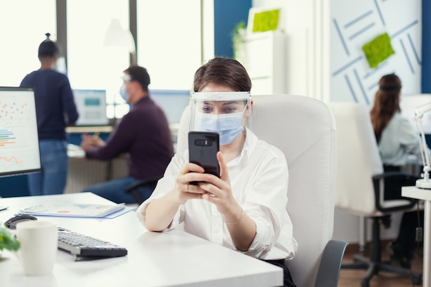 Empresaria charlando en el teléfono inteligente sentado en su lugar de trabajo en la oficina ocupada con mascarilla durante la pandemia mundial con covid19. Compañeros de trabajo multiétnicos que trabajan respetando la distancia social en financia