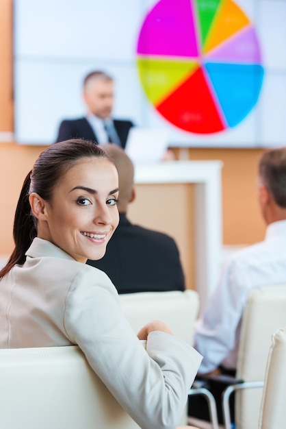 Empresaria en la conferencia. Mujer joven confiada en ropa formal mirando por encima del hombro y sonriendo mientras está sentado en la sala de conferencias con un orador público en el fondo