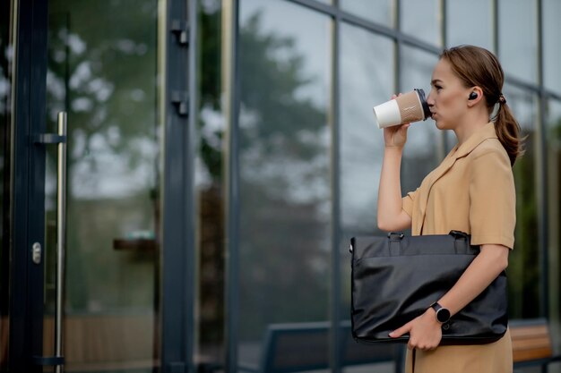 Empresária confiante andando pela cidade falando ao telefone com um fone de ouvido sem fio e relógio inteligente Jovem mulher falando ao telefone e tomando café enquanto caminha