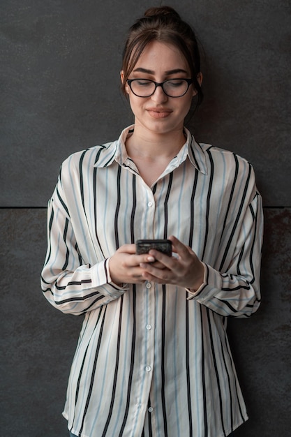 Empresária de inicialização na camisa com um óculos usando smartphone em frente a parede cinza durante uma pausa do trabalho lá fora. Foto de alta qualidade