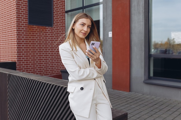 Empresária de terno branco usando telefone celular em pé perto de um edifício moderno