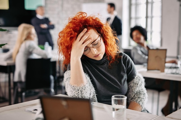 Empresária deprimida segurando a cabeça com dor e se sentindo deprimida no trabalho há pessoas no fundo