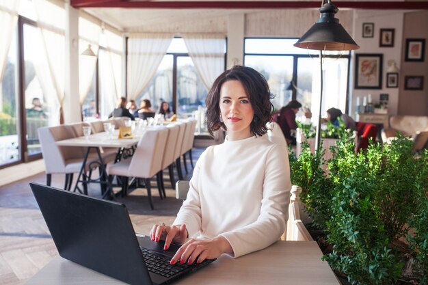 Empresaria elegante adulta en el vestido blanco que se sienta con la computadora portátil en café. Bella dama morena con portátil en café. Foto de una mujer de mediana edad escribiendo en el teclado