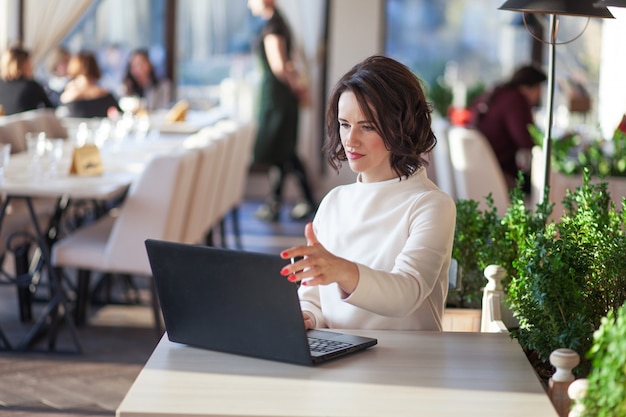 Empresária elegante adulta vestido branco, sentado com o laptop no café. Linda morena usando notebook no café. Foto de uma mulher de meia idade, digitando no teclado