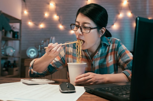 empresária elegante comendo macarrão instantâneo na mesa de jantar enquanto trabalha no projeto com computador portátil e calculadora. fêmea coreana asiática com fome em copos com lanches na hora de dormir à noite.