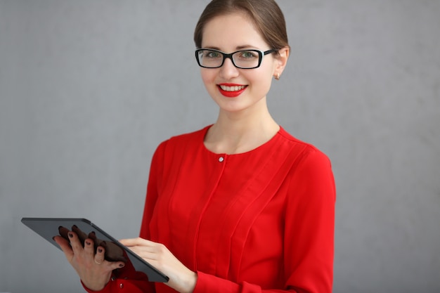 Empresária em uma camisa vermelha e óculos, segurando um tablet nas mãos, sobre um fundo cinza