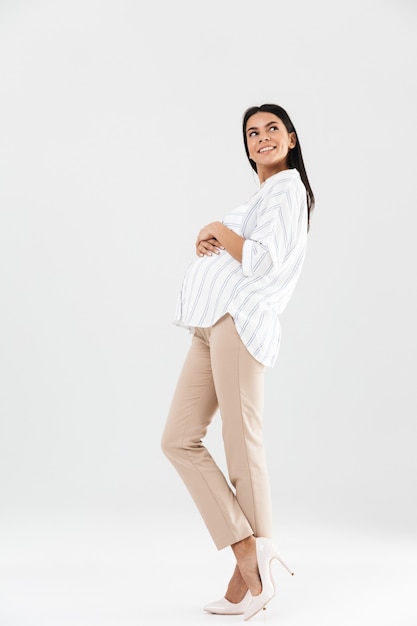Empresaria embarazada europea de longitud completa sonriendo y tocando su gran barriga mientras está de pie aislado sobre la pared blanca