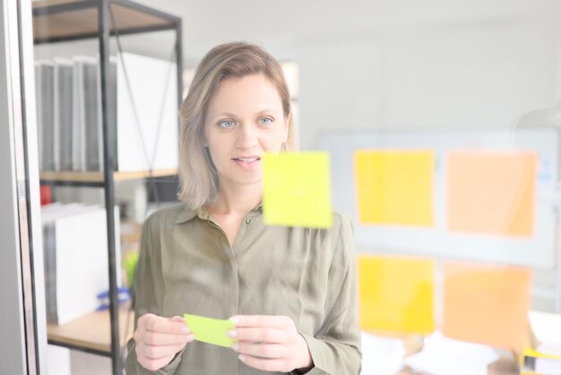 La empresaria se encuentra junto a la pared de vidrio con pegatinas de notas en las hojas de los empleados de la oficina de la empresa