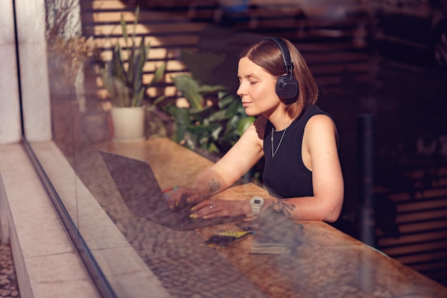 Empresaria enfocada en auriculares que trabajan en una computadora portátil en un acogedor coworking cerca de la ventana