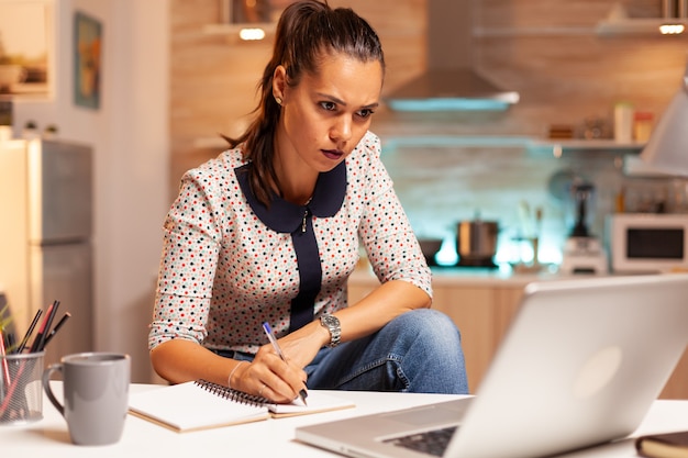 La empresaria escribiendo notas mientras trabaja hasta tarde en la computadora en la cocina de casa. Empleado que usa tecnología moderna a medianoche haciendo horas extras para trabajo, negocios, ocupado, carrera, red, estilo de vida, inalámbrico.