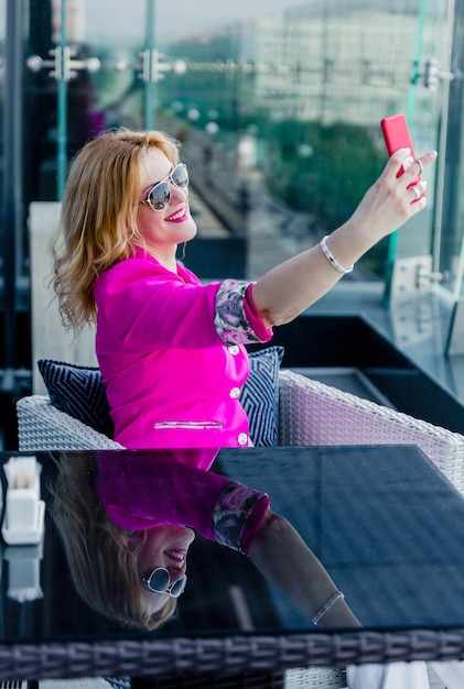 Una empresaria exitosa con una chaqueta magenta se hace un selfie en la terraza de un café en un centro de negocios. Concepto de representación de mujer de negocios