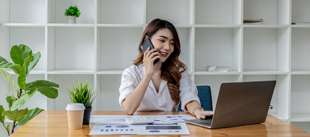 Empresária falando ao telefone com seu parceiro de negócios, ela se senta em uma sala privada, em uma sala decorada com plantas e estantes sobre a mesa com papéis de trabalho e laptop. Conceito de mulheres asiáticas.