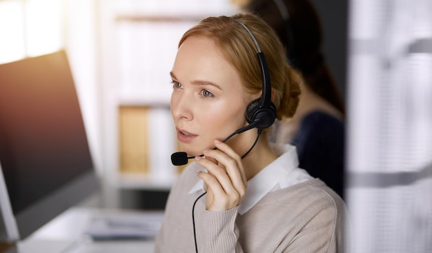 Empresária falando por fone de ouvido enquanto está sentado no escritório ensolarado Call center e grupo de pessoas diversas nos negócios