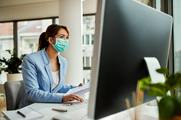 Empresária feminina usando máscara facial enquanto analisa relatórios de negócios e trabalha em PC desktop no escritório