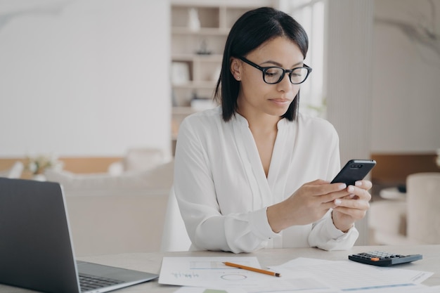 Empresária focada em óculos responde à mensagem do cliente usando aplicativos de celular sentados no laptop