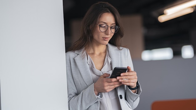 Empresária focada usando óculos com celular na mão em pé no escritório moderno