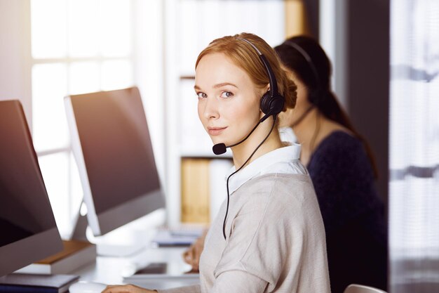 Empresaria hablando por auriculares en la oficina soleada. Centro de llamadas y grupo de personas diversas en los negocios.