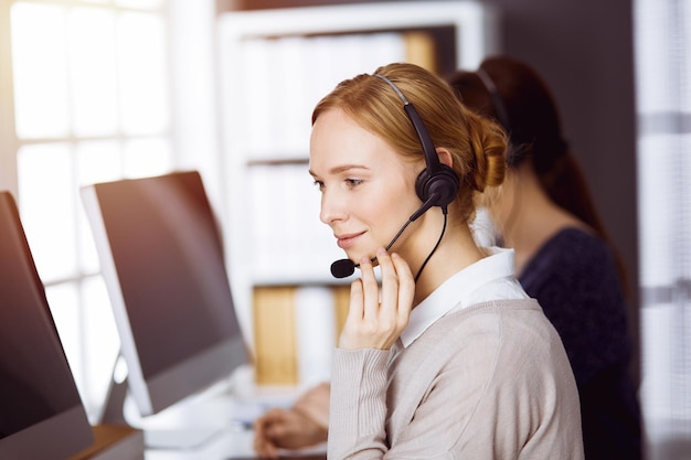 Empresaria hablando por auriculares en la oficina soleada. Centro de llamadas y grupo de personas diversas en los negocios.