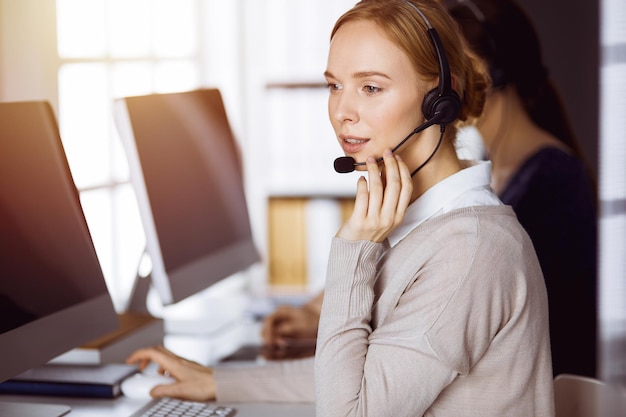 Empresaria hablando por auriculares en la oficina soleada. Centro de llamadas y grupo de personas diversas en los negocios.