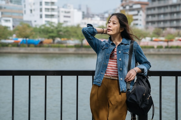 empresária japonesa relaxante perto do rio limpo com ventos soprando o cabelo de férias. jovem escritório descanso contando com corrimão depois do trabalho, apreciando a bela vista da cidade. estilo de vida da população local.