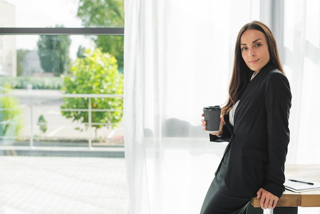 Empresaria joven que se sienta al borde del escritorio de madera que sostiene la taza de café disponible cerca de la ventana