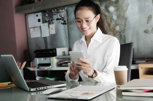 Empresaria joven que usa el teléfono elegante mientras que trabaja en su fondo del escritorio de oficina