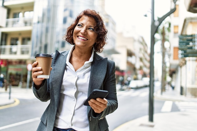 Empresária latina de meia-idade bebendo café e usando smartphone na cidade.